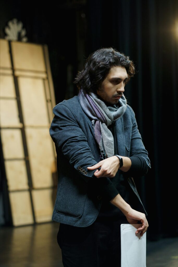 A male actor thoughtfully preparing for a rehearsal on stage, in a dimly lit theater setting.